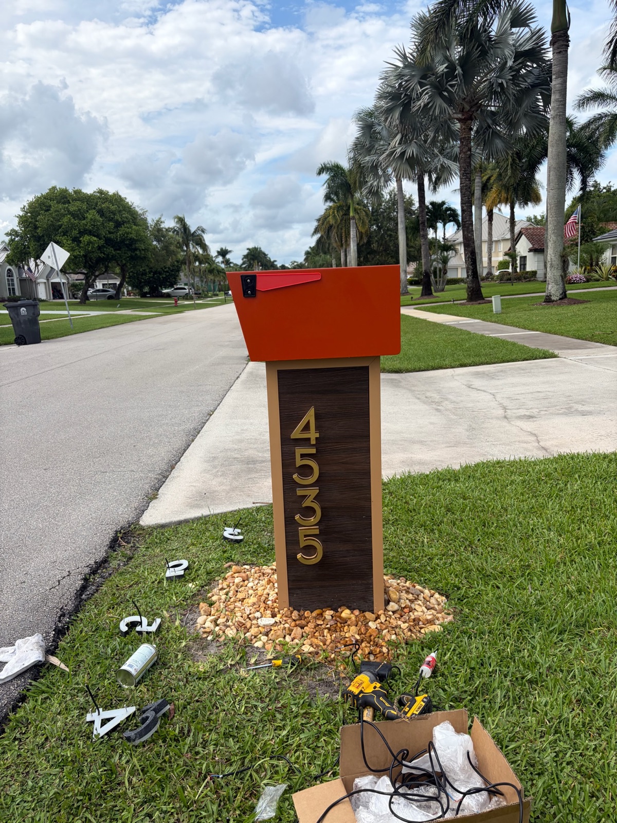 A custom mailbox and post at the curb of an established community, photographed from the street with mature landscaping behind it