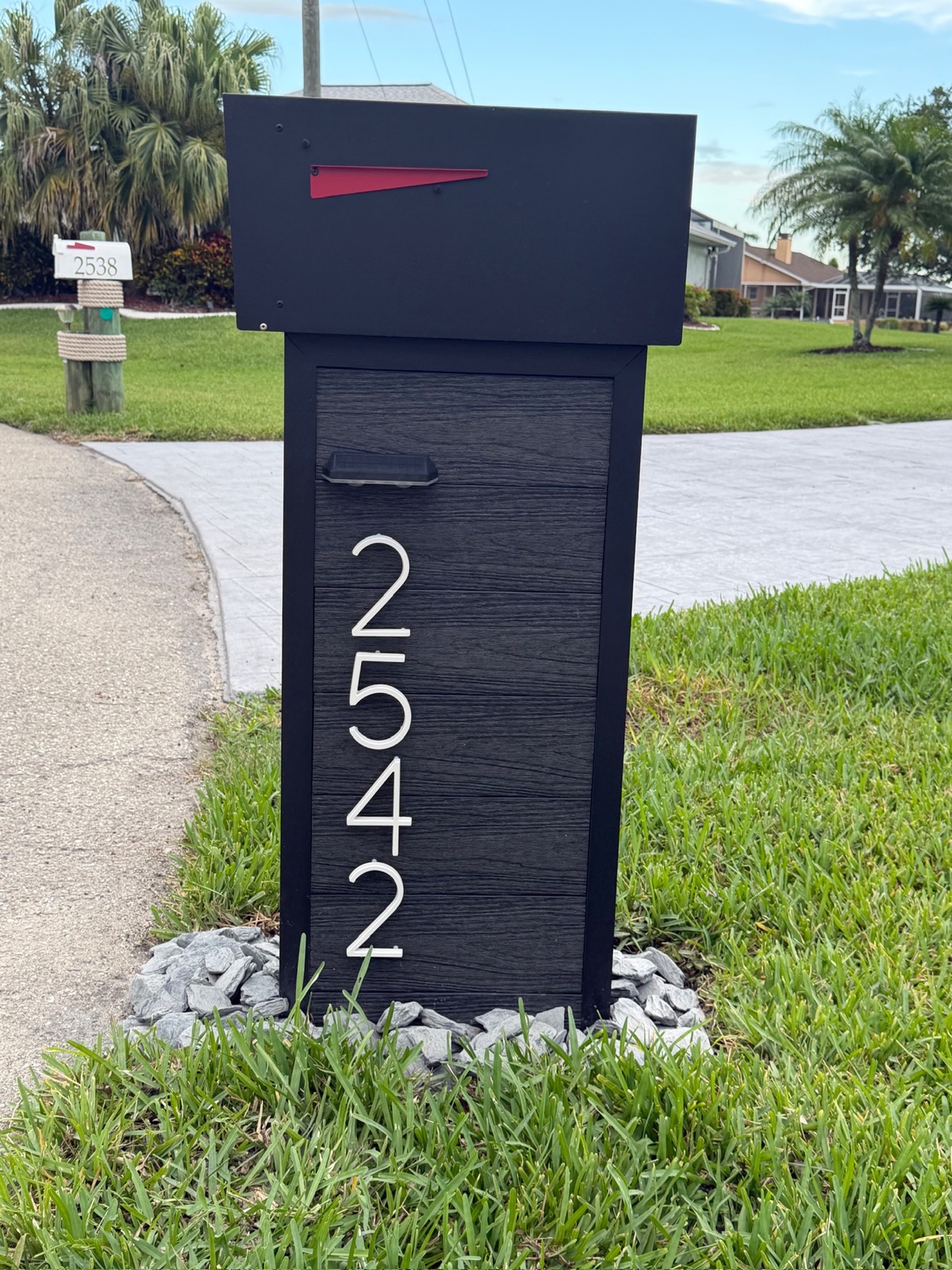 A black composite community mailbox post with vertical white address numbers, set on a gravel bed at the curb